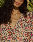 A woman with long curly hair wears the natalie martin Amelia Top, a floral v-neck blouse in red, pink, and blue, paired with a wide-brim straw hat, standing in sunlight among palm leaves.