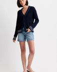 A woman with short dark hair and a neutral expression stands against a plain background wearing the One Grey Day Chelsea Cardigan in navy blue with white sleeve stripes, paired with denim shorts and beige flip-flops.