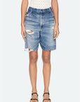 A person wears a sleeveless patterned top and Sea New York’s Milene Denim Knee Short, featuring a relaxed fit and frayed hems, paired with black sandals against a plain light background.