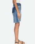 A woman stands in profile wearing a sleeveless white top, tan flat sandals with black straps, and the Milene Denim Knee Short by Sea New York—a knee-length vintage patchwork skirt featuring frayed hem and distressed details.