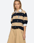 A woman wears the Sea New York Juliet SS Rugby—a puff-sleeve top with beige and navy stripes plus a white collar—paired with a beige skirt and braided brown belt, standing against a plain light background.