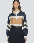 A woman wears the Sea New York Juliet Polo, featuring bold navy, gold, cream, and green stripes, paired with a high-waisted pleated black skirt and belt. She stands against a plain light background.