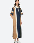 A woman wears the Sea New York Juliet Polo Maxi Dress, a loose-fitting, short-sleeve style with vertical navy, tan, cream, and green stripes. She stands with one hand in her pocket and white shoes against a plain background.