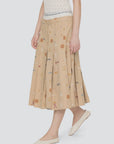 A person wears the Sea New York Juliet Skirt—a beige pleated embroidered skirt with colorful symbols and an athletic waistband—paired with a white lace top and white ballet flats, standing against a plain light background.
