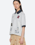 A young woman with straight dark hair wears the Winslow Craft Polo Shirt by Sea New York, a relaxed-fit white top with black stripes and embroidered animals, paired with a tan printed skirt. She stands against a plain light background, looking slightly to the side.