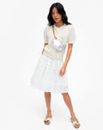 Woman wearing a white short-sleeve cardigan, white skirt, white slide sandals, and the Clare Vivier Petite Fanny S26 silver crossbody bag with adjustable strap, standing against a plain white background.