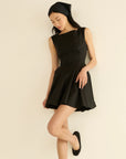 A woman in a sleeveless DOEN Riviette Dress, black flats, and a black headscarf leans gracefully against a light wall, gazing down with one leg slightly bent.