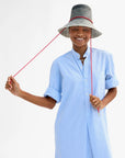 A person wearing a blue and white striped dress smiles while holding the sail cord strap, modeling the Clare Vivier Romy Hat—a black and white wide-brimmed straw rattan hat (57 cm circumference)—against a plain white background.