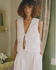 A person with curly, shoulder-length hair wears the Sea New York Lois Twill Vest, styled with loose white pants, standing indoors near a floral curtain and a table with white lilies.