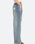 Wearing Sea New York's Claira Mended Jeans—relaxed fit, light blue denim with patchwork and a back pocket label—paired with a white textured top and black sandals, the person stands in side profile.