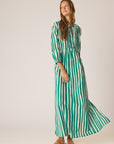 A woman poses in the Sammie Maxi Dress by natalie martin—a green and white vertically striped 100% cotton maxi with three-quarter sleeves and a v-neckline—paired with brown sandals, set against a plain light background.