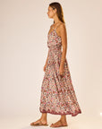 A woman stands in profile wearing the natalie martin Bella Skirt, an ankle-length floral rayon skirt with an elastic waist, paired with flat sandals. She has long hair and poses against a plain, light-colored background.