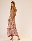 A woman wears the natalie martin Bella Skirt, a long floral-patterned rayon skirt with an elastic waist, paired with sandals. She stands on a plain background, looking back over her shoulder with a neutral expression.