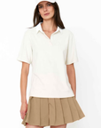 A young woman wearing a Kule cotton terry polo shirt and a khaki pleated skirt with a white cap, standing against a plain white background. She appears confident and casually stylish.