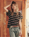A person with long, wavy hair is standing in front of a wooden door. They are wearing a short-sleeved The Boxy Crew by The Great Inc. and high-waisted jeans, with one hand resting on their head and the other in their pocket. The background includes part of a boat's interior.