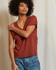 A person with curly hair is standing against a plain background. They are wearing a Hendrix Boxy V-Neck Tee by Perfectwhitetee in a reddish-brown shade, with rolled-up sleeves, paired with blue jeans. The 100% cotton shirt is a staple piece in their wardrobe. The person has one arm crossed over to touch the opposite shoulder and they are looking slightly to the side with a relaxed expression.