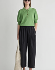 A person wearing a green collared knit top, Apiece Apart Spa Pleat Pant in black, black sandals, sunglasses, a black headband, and holding a cream-colored net tote bag stands with hands in pockets.