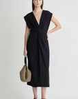 A woman wears the Apiece Apart Margit Twist Dress, featuring a sleeveless deep V-neck with front twist detail, paired with black sandals, a black headband, and a two-handle woven bag against a light background.