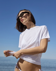 A woman in sunglasses and a white bikini bottom stands by the sea under a clear sky, pulling slightly on her Sold Out NYC Iconically Soft Perfect Tee in classic crew-neck organic Peruvian cotton, and looking off to the side.