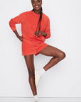 A Black woman with braided hair wearing a Kule Terry Franny sweatshirt and shorts set in orange, along with white sneakers, playfully poses with one leg lifted against a white background.