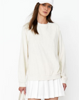 A woman in a stylish white outfit including a cap, Kule's Terry Franny sweatshirt, and pleated skirt, paired with a shoulder bag, posing against a plain background.