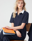 A woman with blond hair sits on a wooden stool, holding an orange notebook. She wears a machine washable, navy blue Kule terry polo shirt with a white collar and looks thoughtfully at the camera.