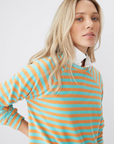 A person with long blonde hair is posing against a plain background. They are wearing The Terry Franny sweatshirt by Kule, which is a cotton-blend, striped sweater with teal and orange horizontal stripes over a white collared shirt. The person has a neutral expression, and their head is slightly tilted.