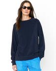 A woman with long, dark hair wearing black sunglasses and a navy blue Kule The Terry Franny sweatshirt with raglan sleeves. Her bright blue pants contrast the plain white background as she stands confidently with her hands in her pockets.
