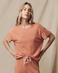 A person with long blonde hair stands confidently with hands on hips. They are wearing a matching, 100% cotton peach-colored outfit with a relaxed fit, featuring The Boxy Crew by The Great Inc., a short-sleeved top, and pants that feature a drawstring. The background is a neutral beige canvas.