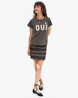 A person with long dark hair stands against a plain background. They are wearing a pre-shrunk, 100% cotton T-shirt with the word "OUI" printed on it, specifically the Classic Tee - OUI by Clare Vivier, and a patterned skirt. They have black ballet flats with ankle straps and are accessorized with a necklace and a bracelet.