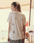 A person with light brown hair stands on a sunny deck facing away from the camera. They are wearing The Boxy Crew by The Great Inc., a white T-shirt with various colorful embroidered designs, including a heart, rainbow, star, and playing cards—reminiscent of vintage tees. They also have leopard-print shorts.