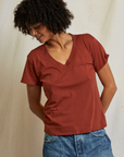 A person with curly hair is standing against a light background, looking down and to the side while smiling. They are wearing a rust-colored Hendrix Boxy V-Neck Tee from Perfectwhitetee and blue jeans, an essential staple, with hands held behind their back.