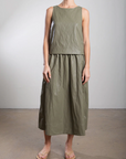 Wearing Pharaoh's Claudia Skirt and a sleeveless olive green top, the person stands against a light background. The chic ensemble, made of textured fabric with matching midi skirt and elastic waist, is paired with sandals and crafted in Los Angeles.