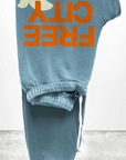 Folded blue FREECITY Large Sweatpant by Free City (sparrow, LLC), made from lightweight French terry, features an orange "FREE CITY" print and partial white graphic at the top left. Includes a white drawstring, placed on textured concrete against a plain background.
