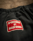 A close-up of dark fabric shows a stitched patch with "SPONSORED BY MADEWORN" in white on red. The heavyweight French terry cotton implies these could be MadeWorn P.E. Sweatpants, meticulously handmade in Los Angeles and potentially unisex.