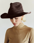 A person with short blonde hair wears the Janessa Leone Sacha Hat, gold hoop earrings, and a beige turtleneck top, looking downward against a plain light background.