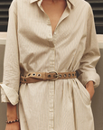 Xirena Boden beige striped cotton dress with rolled sleeves and tan grommet belt, model’s hands in pockets.