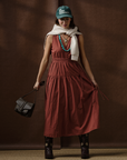 A woman stands before a brown backdrop wearing the Xirena Meri Dress in rust, dark cowboy boots, a white sweater over her shoulders, a green "LIL COWBOY" hat, turquoise necklace, and holding a small black bag.