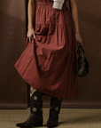 Wearing the Xirena Meri Dress in rust, paired with black floral cowboy boots and a small dark handbag, this look features soft cotton fabric for comfort. The brown background and unseen face keep the focus on the outfit.