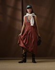 A woman stands indoors wearing the rust-colored Xirena Meri Dress, black cowboy boots with stars, a green hat, turquoise necklaces, a white sweater draped over her shoulders, and carries a small black bag. Brown fabric forms the backdrop.