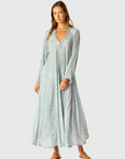 A woman stands barefoot against a plain light gray background, modeling the natalie martin Fiore Maxi—a long sleeve, flowing A-line dress in a light blue pattern with a v-neckline.