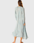 A woman with long hair stands with her back to the camera in a natalie martin Fiore Maxi—a loose, light blue patterned A-line dress with a v-neckline and long sleeves—paired with sandals against a plain background.