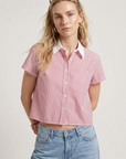 A woman with long blonde hair and sunglasses models THE SHIRT’s Boxy Short Sleeve Cropped Shirt—red and white striped, featuring No Gape button technology—paired with light blue jeans against a plain background.