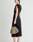 A woman stands sideways against a plain light background wearing the Apiece Apart Margit Twist Dress in black cotton jersey, paired with black sandals, a black headband, and holding a woven dome-shaped handbag.