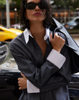 A woman with wet, chin-length hair and dark sunglasses wears The Everything Shirt by Sold Out NYC—an oversized pinstripe style with crisp white cuffs and collar—carrying a jacket with a blurred yellow car in the background.