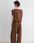 A person with short curly hair stands sideways, wearing the Rachel Comey Rivera Top—a relaxed crinkled blouse with wide flutter sleeves—paired with matching pants, against a plain light background.