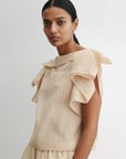 A woman with long dark hair tied back wears the Rachel Comey Rivera Top, an airy beige blouse in crinkled fabric with large ruffled flutter sleeves. She stands against a plain light background, looking calmly at the camera.