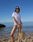 A woman in sunglasses, The Iconically Soft Perfect Tee by Sold Out NYC, and bikini bottoms stands on a rocky beach with a woven straw bag, ocean waves, and blue sky in the background.