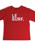 The Le Surf Tee by Ping Pong Surf Club is a loose-fit red T-shirt made from soft combed cotton, featuring "LE SURF." in bold white capital letters across the chest.