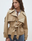 A woman with wavy brown hair models the SUNCOO Elliott Jacket over a brown and white polka dot collared shirt and blue jeans, standing against a plain light background and looking to the side.